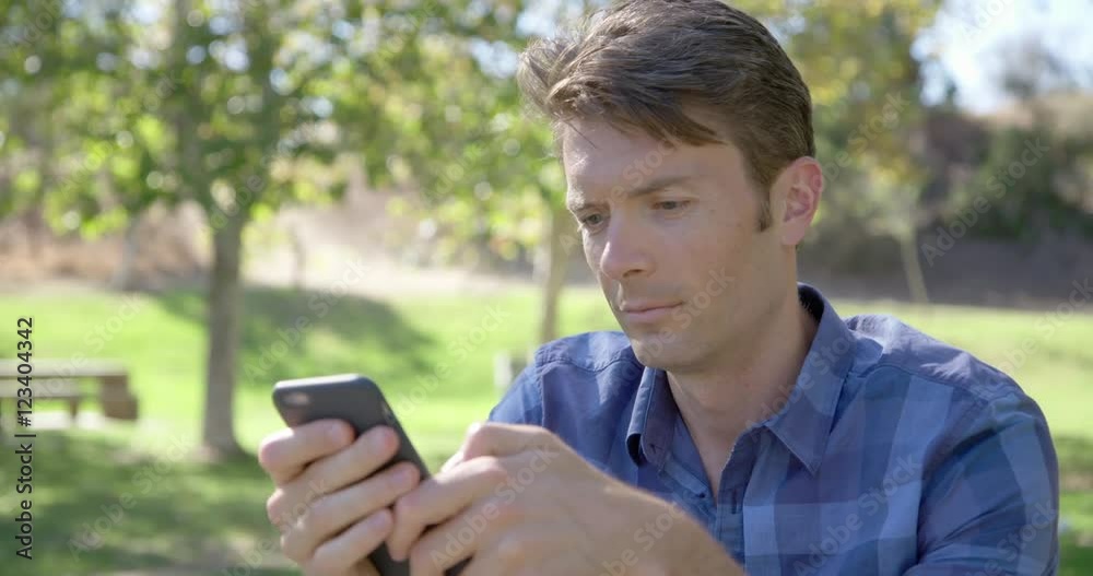 Young man sitting in a park, texting and surfing the internet on a smart phone on a sunny day in Los Angeles. Close up, recorded hand-held in real time from front. Young man sitting in a park, texting and surfing the internet on a smart phone on a sunny day in Los Angeles. Close up, recorded hand-held in real time from front.