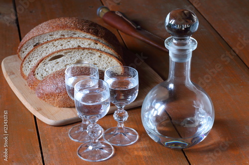 Графин и три рюмки с водкой стоят на деревянном столе рядом с ломтями хлеба.
