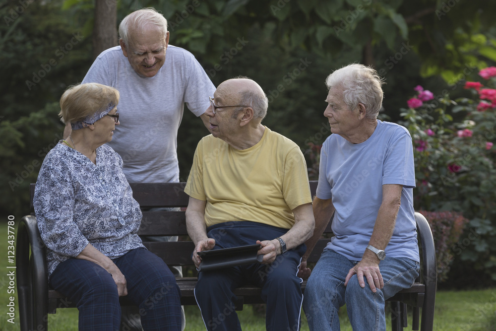 Smiling senior friends talking while using digital tablet at park bench Smiling senior friends talking while using digital tablet at park bench