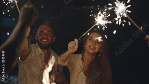 Young people celebrating something on the beach at night with firework sparklers, one couple kissing and hugging