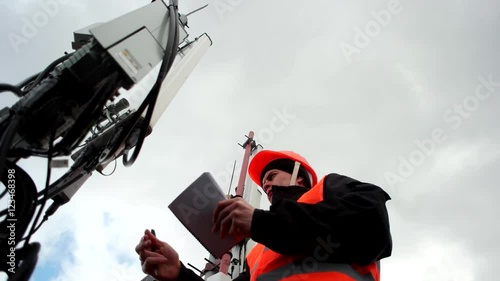 employees mobile Company, cellular, an engineer working with the antennas and transmitters on the tower, records data on a background of gray sky, a man dressed in uniform and hel photo