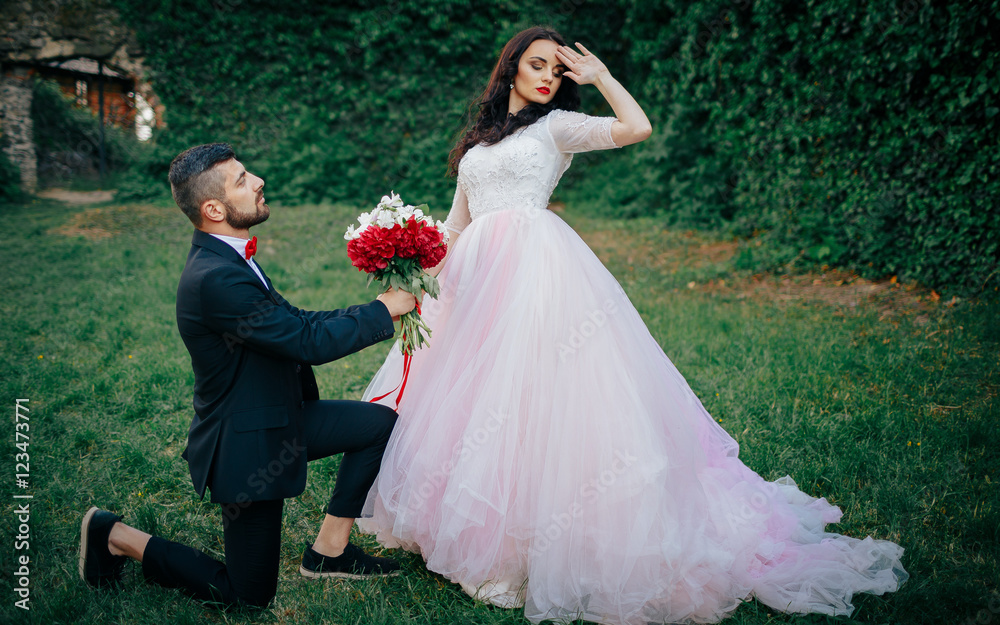 Stylish bearded groom in a tuxedo with a red butterfly and suspenders with his bride with long black hair in a pink long dress is holding a bouquet of peonies with a tied ribbon walking in green park