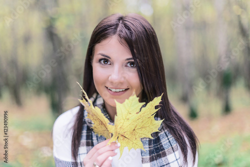 A young  smiling girl in a white knit scarf is holding yellow maple leaves on the background autumn forest