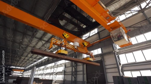 Yellow overhead crane with electromagnetic beam grippers carries steel cargo in engineering plant shop warehouse.