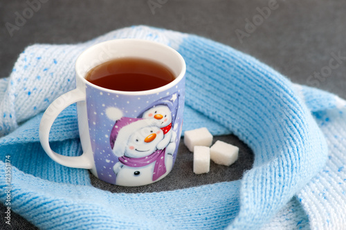 Кружка чая с рисунком снеговика и снега. Голубой вязаный шарф окутывает кружку чая, создавая обстановку домашнего уюта. Кубики сахара лежат рядом с кружкой чая.