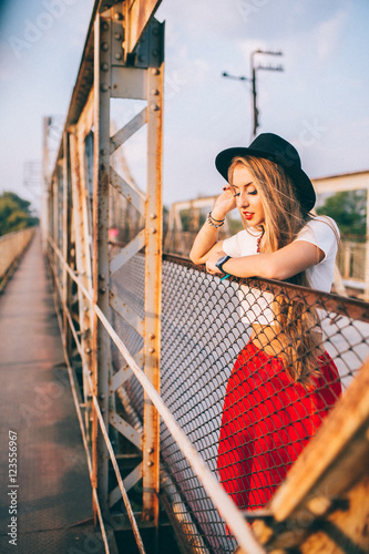 stylish portrait of a beautiful young woman on the bridge in a red skirt and black hat on a sunny day at sunset posing and smiling Life Style