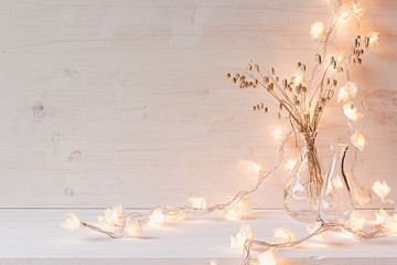 Burning fairy lights and dried spikelets in glass vase on white wooden background, soft warm cozy winter interior decoration with copy space.