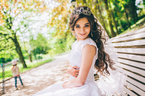 fashion outdoor photo of gorgeous bride with dark hair in luxurious wedding dress and crown posing at the park. close up. gemstone