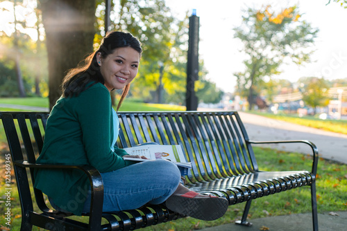 College aged girl smiling and reading a book on a bench in a park
