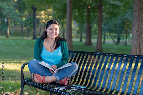 Smiling female college student reading a book on a bench in a park