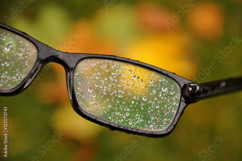 Glasses in a black frame with raindrops on the lens close-up. Blurred autumn background.