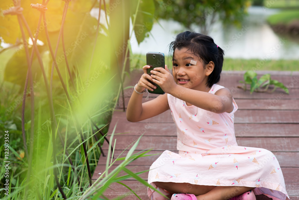 Adorable little girl taking a photo by smartphone against bokeh tree background,she is confused about photography Adorable little girl taking a photo by smartphone against bokeh tree background,she is confused about photography