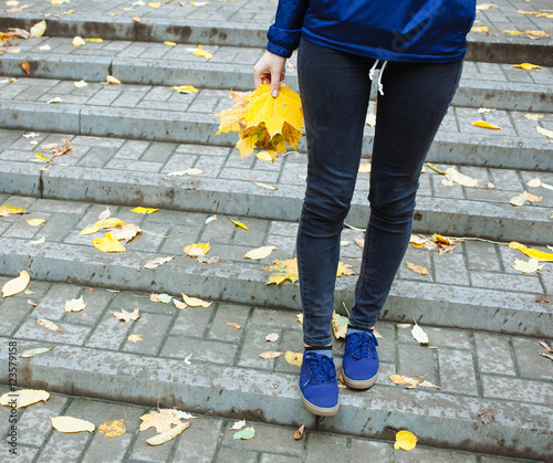Part of body people cold rainy autumn concept. Sporty legs blue jeans and sneakers. Woman in blue wind braker holding leaves in hand.