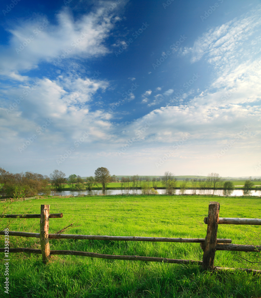 Spring Landscape of Green Meadow with Wooden Fence along River under Blue Sky