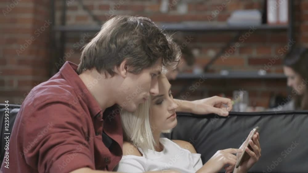 Attractive young couple watching smart phone together sitting on a couch in a living room at modern loft interior. Beautiful young hipster couple using phone at home. Attractive young couple watching smart phone together sitting on a couch in a living room at modern loft interior. Beautiful young hipster couple using phone at home.