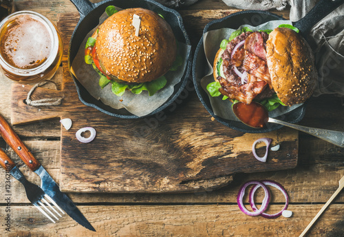 Homemade beef burgers with crispy bacon and fresh vegetables in small pans and glass of wheat beer on rustic serving board over shabby wooden background, top view, copy space, horizontal composition