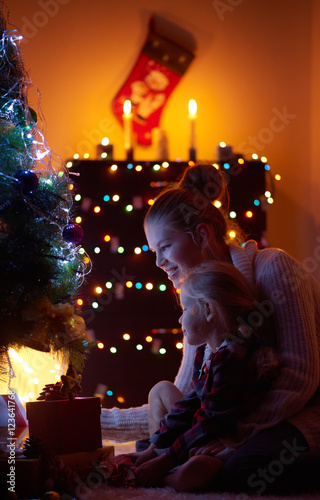 New year fairy tale. Young smiling woman sitting on the floor with little daughter  looking at Christmas tree and lights. Concept of cozy room decoration and presents. 