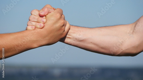 Friendly handshake of two unrecognizable muscular white men on blue sky background. Shaking of male arms outdoor. Two strong men having firm handshake outside. Teamwork and friendship. Close-up © olehslepchenko