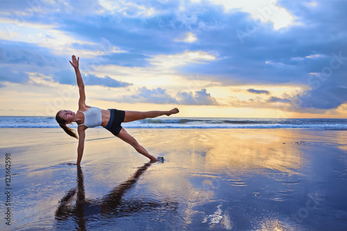 Active sporty woman stand in yoga pose on black sand beach to keep fit and health. Sunset blue ocean surf background with sun. Woman fitness, morning workout, sport activity on summer family holiday