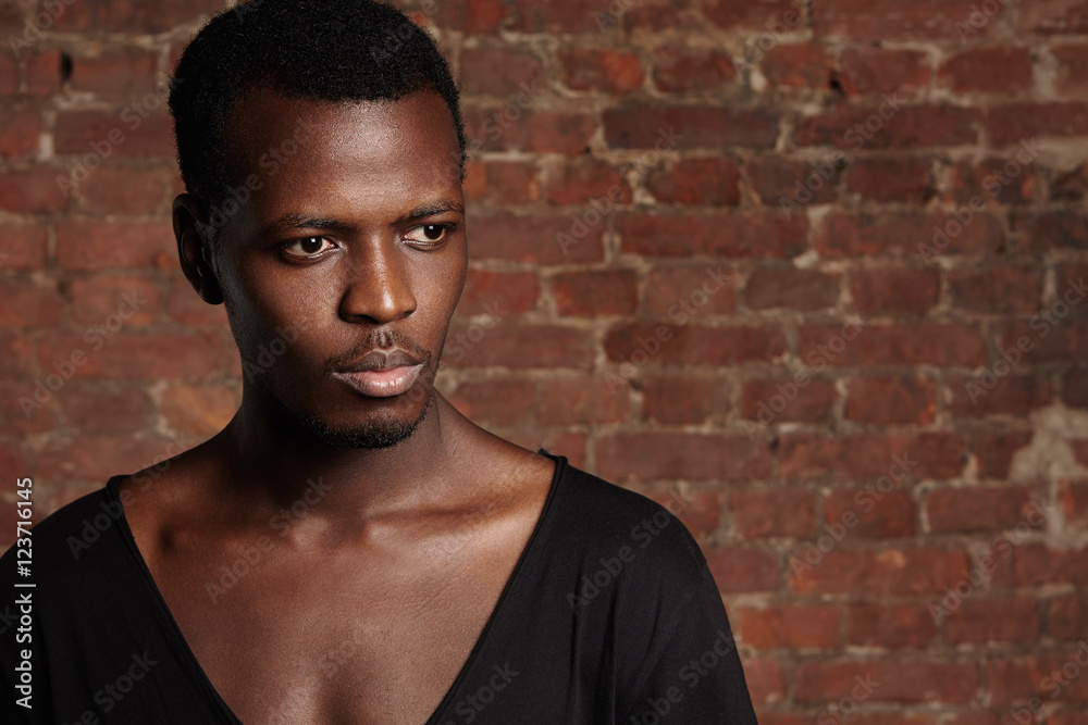 Fashion and beauty concept. Portrait of handsome stylish young dark-skinned man looking confident and serious, posing in studio against brick wall background for your text or promotional content Fashion and beauty concept. Portrait of handsome stylish young dark-skinned man looking confident and serious, posing in studio against brick wall background for your text or promotional content