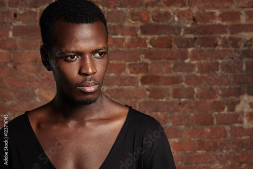 Fashion and beauty concept. Portrait of handsome stylish young dark-skinned man looking confident and serious, posing in studio against brick wall background for your text or promotional content