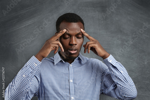 Handsome serious puzzled African student dressed in checkered shirt robbing his forehead, closing his eyes, looking concentrated and focused, trying to remember right answer during test in classroom