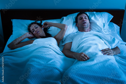 Sleeping man with obstructive sleep apnea snoring loudly in bed while tortured wife plugs her ears photo