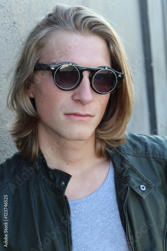 Handsome young man with blond long hair and sunglasses isolated on grey background. Fashion studio shot.