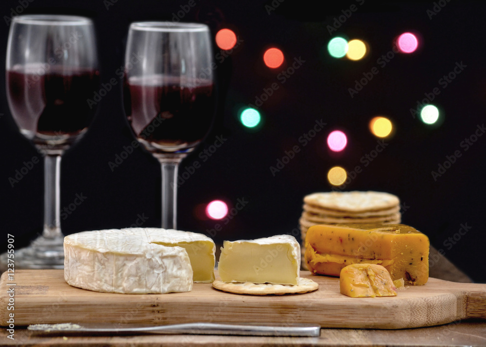 Brie and hot pepper jack cheese on wooden cheese board, accompanied by crackers and two glasses of red wine, against black background decorated with colorful holiday lights Brie and hot pepper jack cheese on wooden cheese board, accompanied by crackers and two glasses of red wine, against black background decorated with colorful holiday lights