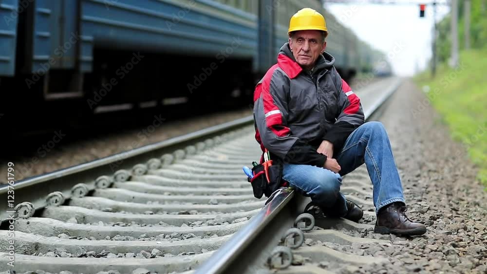 Railway worker sits on railway line. Train rides on the railroad. Railwayman in yellow hard hat sits on rail and looks at the camera. Workman on railway track Railway worker sits on railway line. Train rides on the railroad. Railwayman in yellow hard hat sits on rail and looks at the camera. Workman on railway track