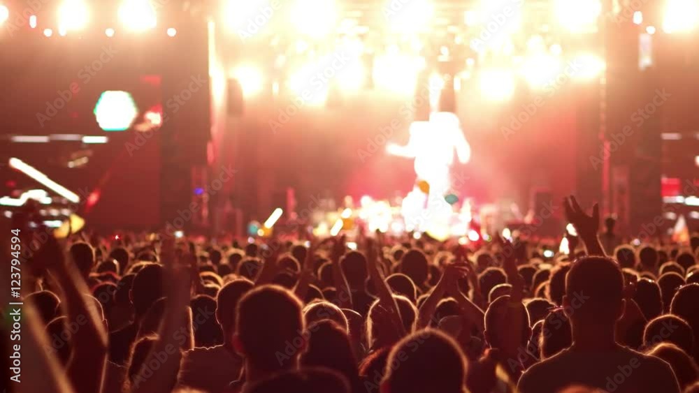 A crowd of people at a concert, a show of hands waving, clap rock musicians on the brightly lit stage, on the big screens in the background can be seen artists, bright multi colored lights on stage