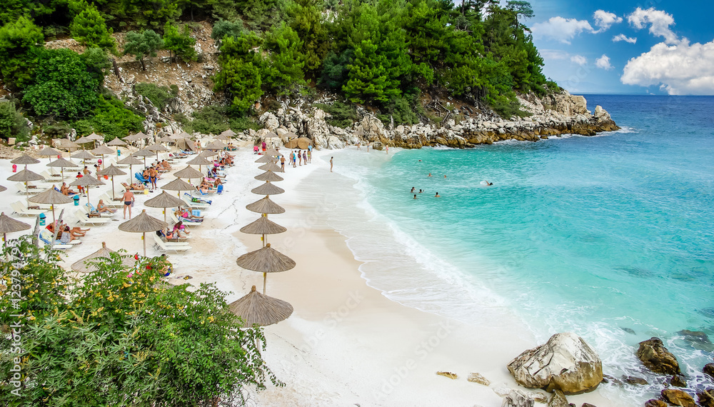 Fototapeta premium Marble beach (Saliara beach), Thassos Islands, Greece. The most beautiful white beach in Greece. Tourists enjoying a nice day at the beach. Straw umbrellas (straw parasol) and sunbeds on the beach.