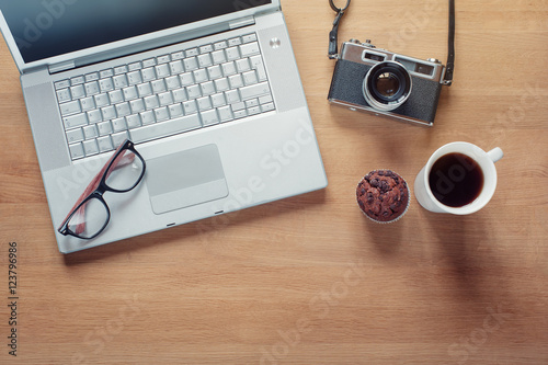  Overhead of modern comfort work place. Different objects on wooden background. Items include camera, glasses, laptop, cup of coffee and muffin