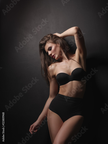 Seductive, beautiful and attractive girl with thick brown hair and sexy gorgeous body is posing in the black seamless lingerie on the dark background in the studio © maksimvostrikov
