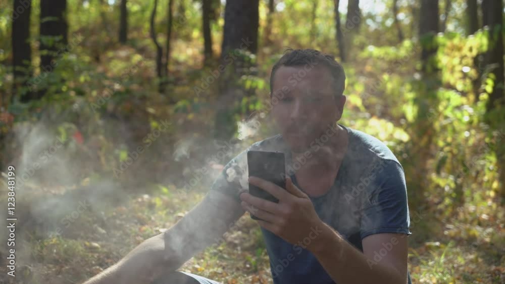 Adult man smoking electronic cigarette in the park or forest. Handsome male wearing in casual t-shirt and jeans sitting on the grass. Guy using smartphone read news or surfing internet or chatting Adult man smoking electronic cigarette in the park or forest. Handsome male wearing in casual t-shirt and jeans sitting on the grass. Guy using smartphone read news or surfing internet or chatting