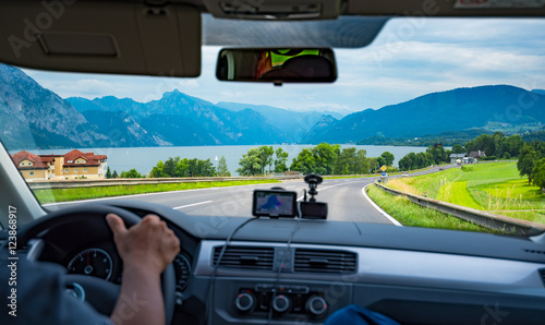 Fototapeta Naklejka Na Ścianę i Meble -  Hands of a driver on steering wheel driving on the road and using GPS with beautiful lake and mountain view in Switzerland , blurred effect on front console
