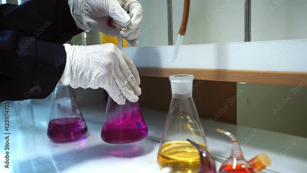 Hands in gloves mixes pink liquid and yellow liquid in flask in lab.