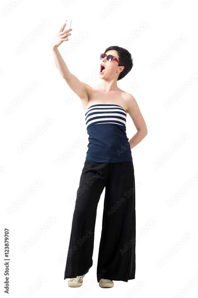 Young girl wearing wide-leg pants and off-the-shoulder top taking expressive selfie. Full body length portrait isolated over white studio background. Young girl wearing wide-leg pants and off-the-shoulder top taking expressive selfie. Full body length portrait isolated over white studio background.