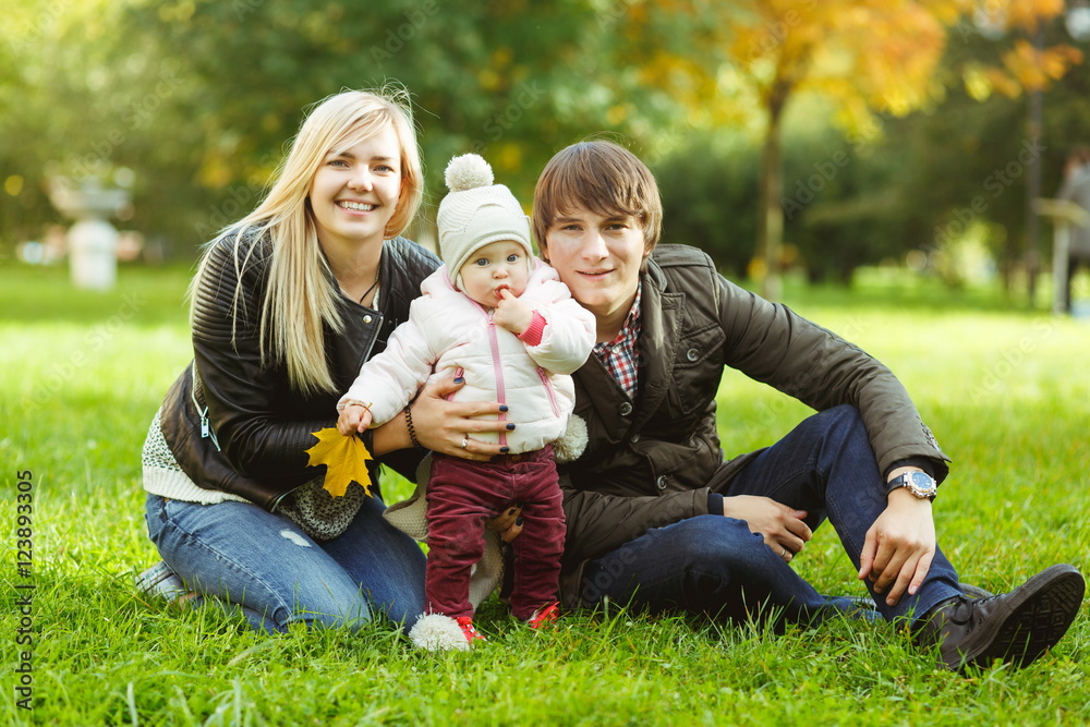 Fototapeta premium Happy parents with daughter sitting on grass in autumn park