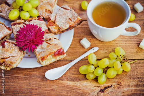 Homemade pie with jam on a plate, cup of tea, grapes, pieces of sugar and aster flower on wooden background. Snack or breakfast serving.