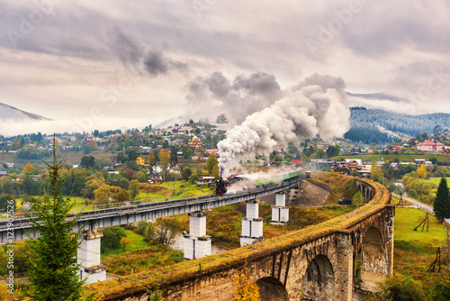 Old railway viaduct in Carpathian mountains with a steam train passing over, beautiful countryside landscape
