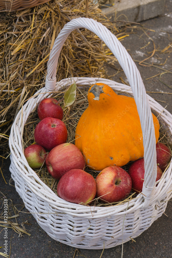 white wicker basket with red apples and orange pumpkin near haystack