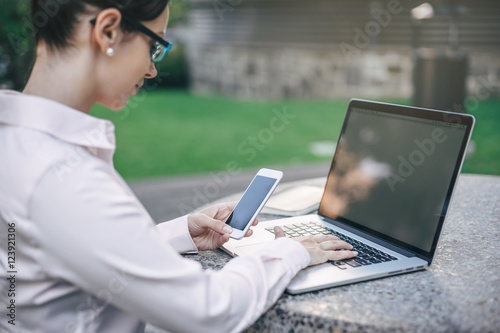 Close-up image of young woman wearing white shirt using smartphone and laptop outside, Business woman checking new messages on cell phone, blurred background, shallow DOF