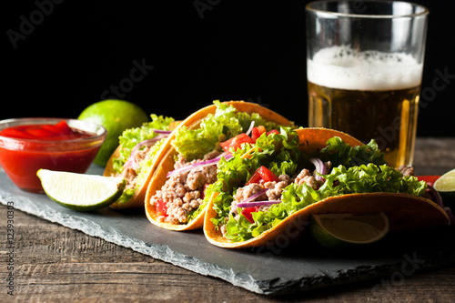 Photo of Mexican tacos with ground meat, beef, beans, onions and salsa on wooden background. Ketchup sauce and lime. A glass o beer in the background.