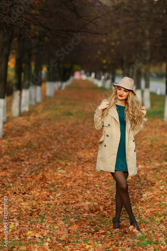 Happy young woman in park on sunny autumn day. Cheerful beautiful girl in coat and floppy hat outdoors on beautiful fall day. Retouched  vibrant colors.