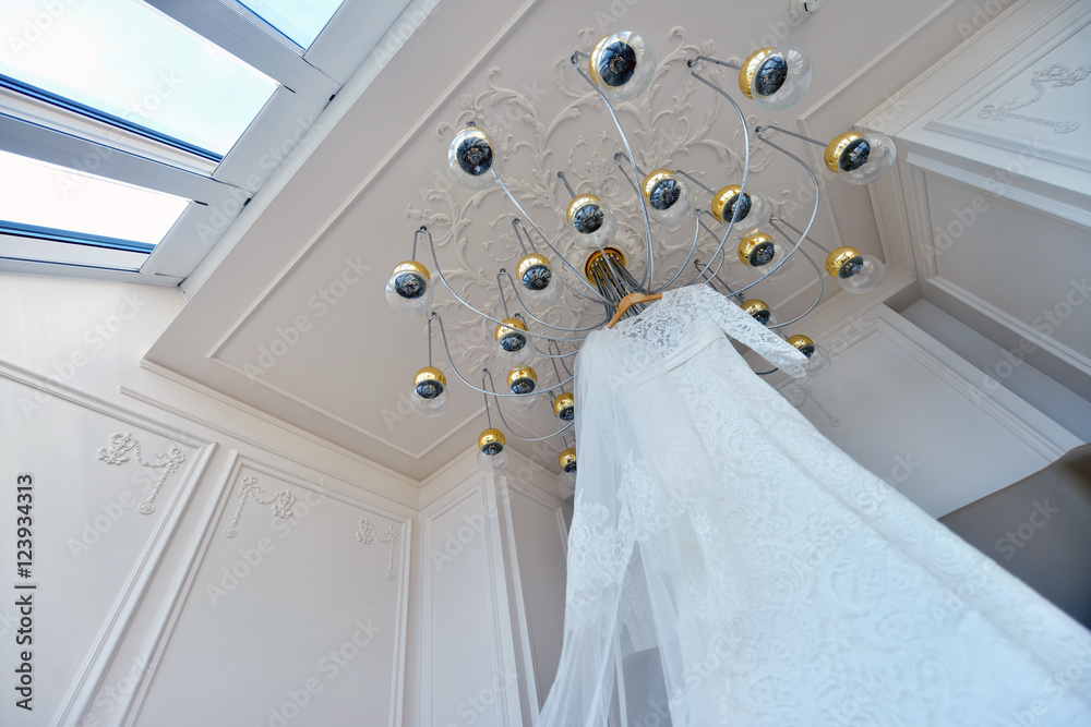 Beautiful white wedding dress for bride indoors on the chandelier. Beauty of bridal gown for marriage. Female lace clothes for celebration Beautiful white wedding dress for bride indoors on the chandelier. Beauty of bridal gown for marriage. Female lace clothes for celebration