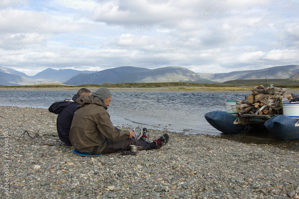 Two men have a rest on a halt, sitting on the pebble coast of the mountain river. Small Usa river, Polar Urals.