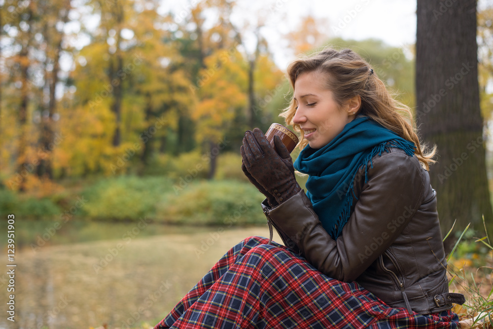 Beautiful girl blonde with wavy hair sitting in the park on autumn leaves and drinking goryady drink from a mug Beautiful girl blonde with wavy hair sitting in the park on autumn leaves and drinking goryady drink from a mug