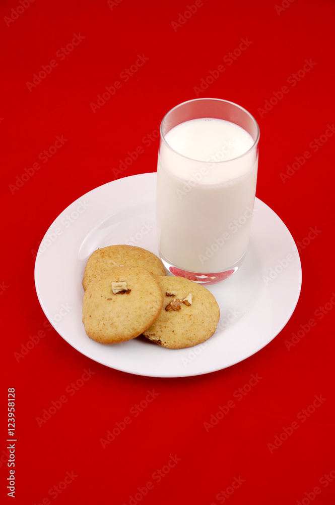 Milk and cookies. A mug of milk and a few home made ginger bread and walnuts cookies on a plate, isolated on red background.Christmas morning. Milk and cookies. A mug of milk and a few home made ginger bread and walnuts cookies on a plate, isolated on red background.Christmas morning.