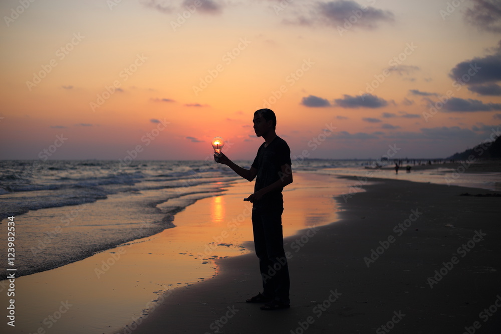 Man holds the electric light bulb and the sun on the beach at sunset